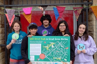 Group of youths holding a board depicting a map of their wild street trails that they have designed. They are stood under a sign saying Community Hub and Library